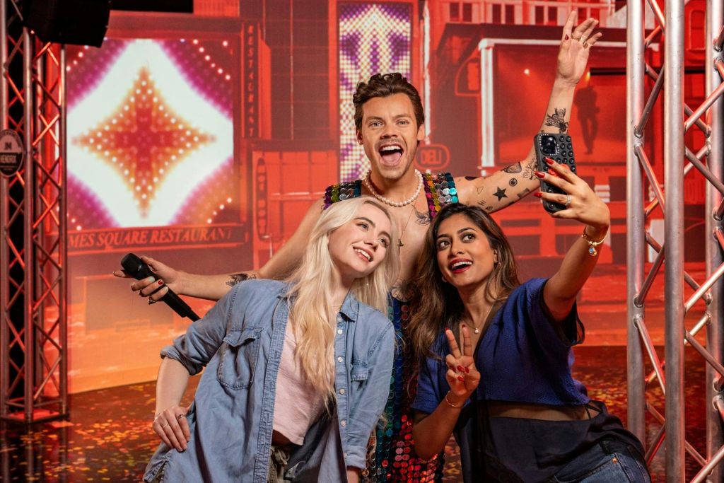 활기차고 밝은 분위기에서 두 여성이 화려한 의상을 입은 남성의 왁스 동상과 함께 미소를 짓고 셀카를 찍으며 포즈를 취하고 있습니다.