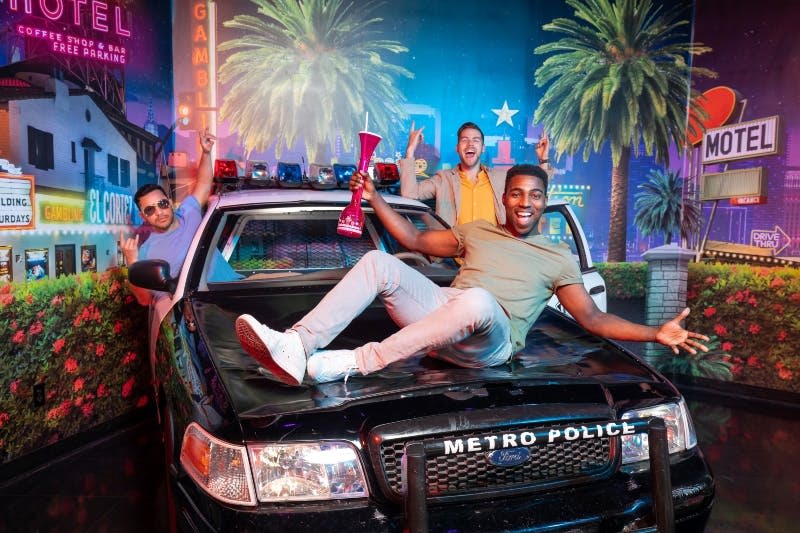 웃고 있는 두 남성이 "Metro Police" 차량에서 포즈를 취하고 있으며, 한 남자는 커다란 음료를 들고 네온 모텔 표지판과 야자수를 배경으로 포즈를 취하고 있습니다.