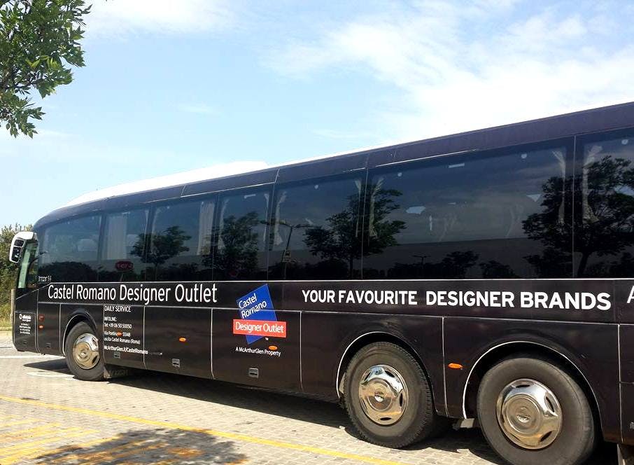 맑은 하늘 아래 포장된 표면에 "Castel Romano Designer Outlet" 브랜드가 새겨진 블랙 투어 버스가 주차되어 있습니다.
