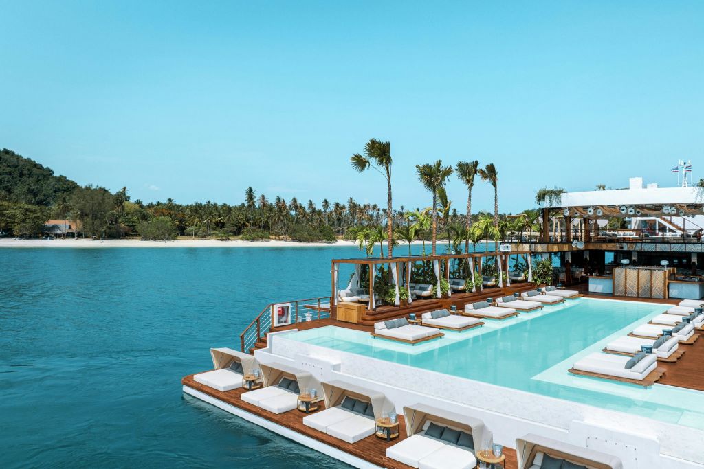 맑고 푸른 물 위로 선착장이 있는 수영장 옆 라운지 공간. 섬을 배경으로 하얀 라운지 의자와 야자수가 있습니다.