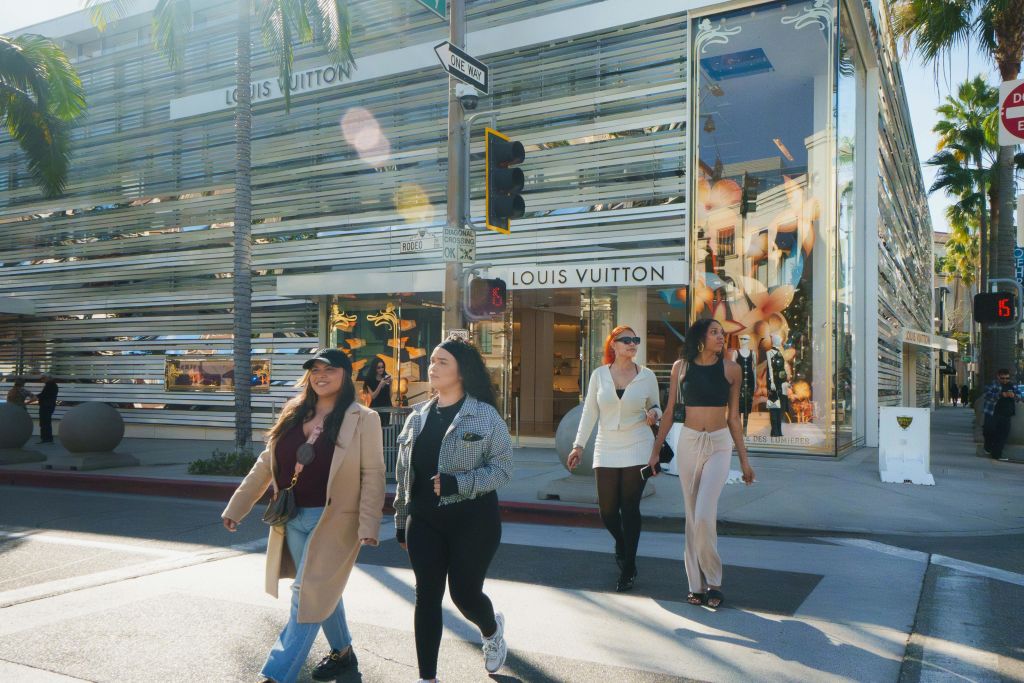 루이비통 매장 앞 햇살 가득한 거리를 가로지르는 네 명의 여성이 야자수와 유리잔에 비친 모습을 감상하고 있습니다.