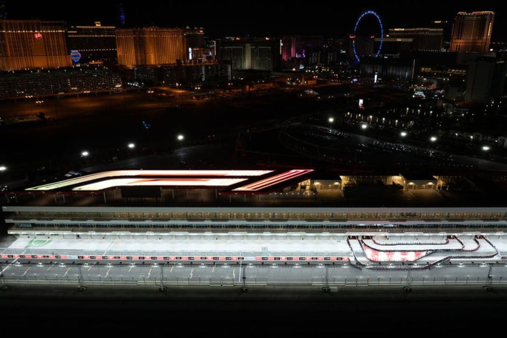 도시의 스카이라인과 대관람차를 배경으로 밝은 피트 레인 조명이 있는 경마장의 야간 공중 전망.