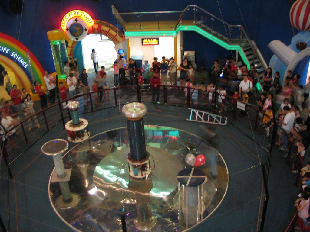 밝게 빛나는 실내 공간에서 난간으로 둘러싸인 높은 원통형 장치로 과학 전시회를 관찰하는 사람들의 군중.