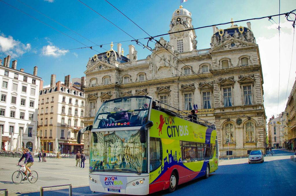 맑고 푸른 하늘 아래 화려한 유서 깊은 건물 앞에 화려한 2층 리옹 시티 버스가 있습니다.