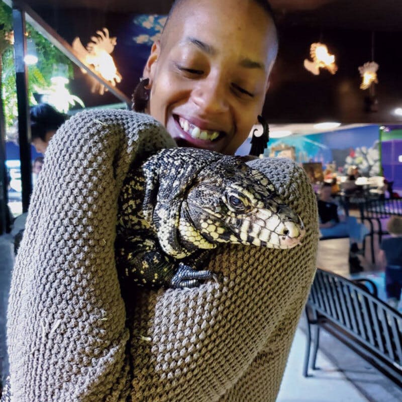 흑백 비늘이 달린 커다란 도마뱀을 안고 웃고 있는 사람. 눈에 띄는 조명과 화려한 벽화를 배경으로 한 실내 환경.