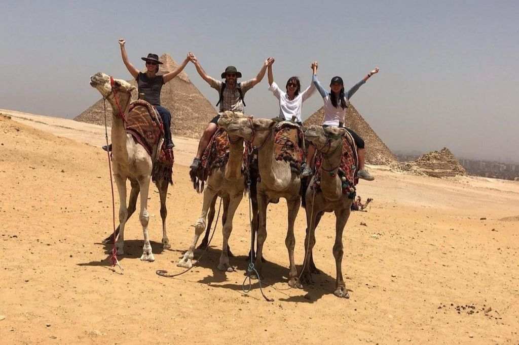 사막에서 두 팔을 들고 낙타를 타고 피라미드 앞에서 손을 잡고 있는 네 사람.
