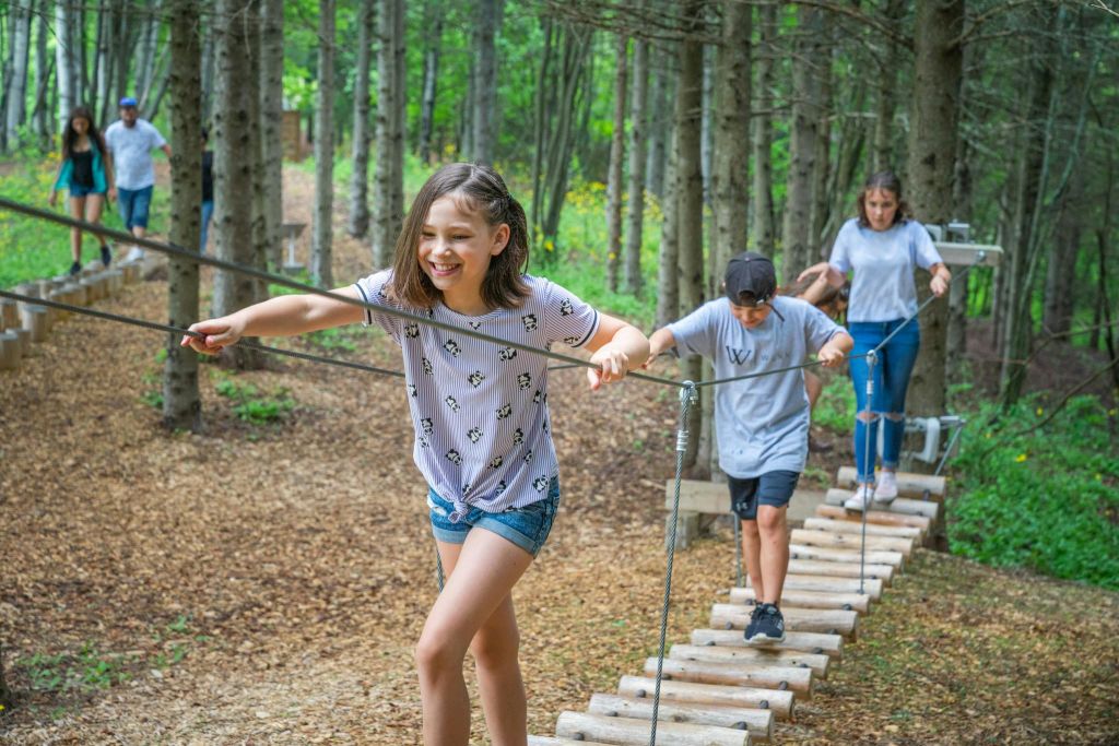 하얀 셔츠와 반바지를 입은 미소 짓는 소녀가 숲의 밧줄 다리에서 균형을 잡습니다.