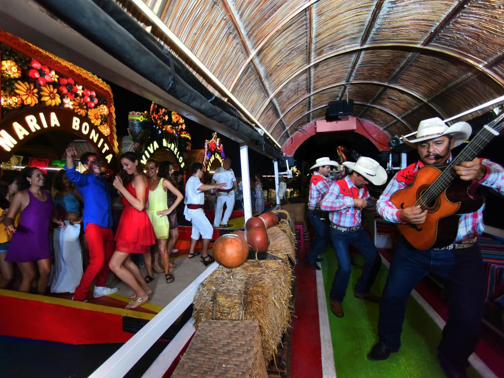 카우보이 모자를 쓴 뮤지션은 초가 지붕 아래 보트에서 기타를 연주하고, 화려한 복장을 한 사람들은 춤을 추며 분위기를 즐깁니다.