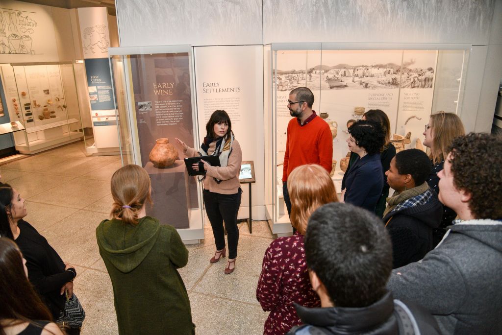 한 무리의 사람들이 전시 케이스와 정보 패널을 배경으로 한 여성이 박물관을 둘러보는 이야기를 듣고 있습니다.