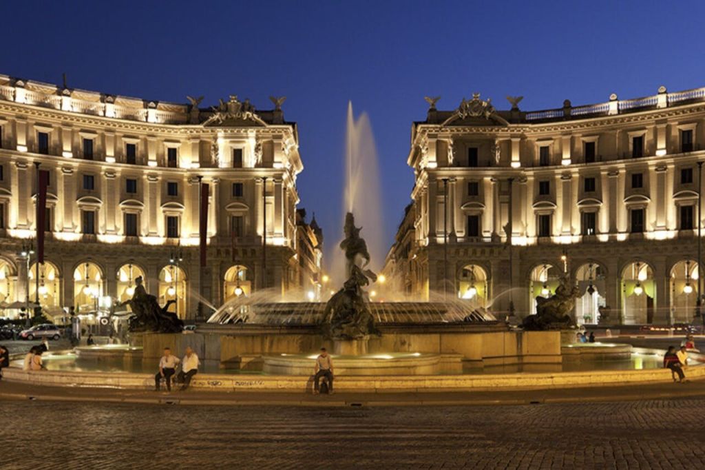 야간에 조명이 켜진 건물과 조각상이 있는 분수대, 인근 공공 광장에 몇 명이 앉아 있습니다.