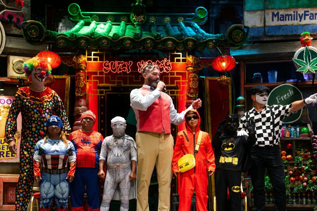 슈퍼히어로와 마임을 포함한 다양한 의상을 입은 사람들이 등불 아래 화려한 무대 위에 서 있습니다.