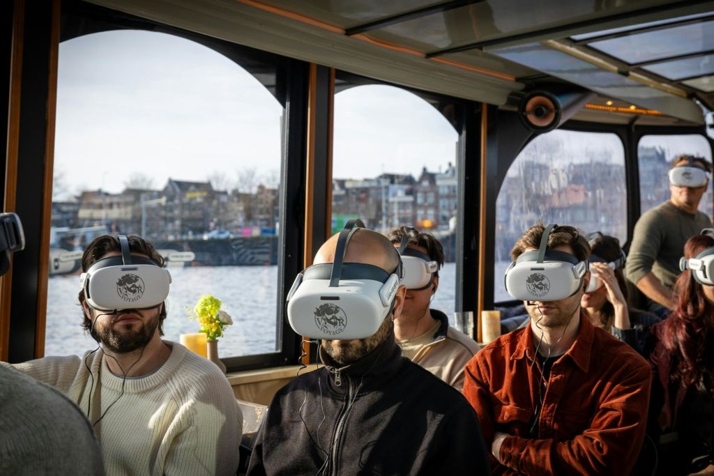 가상 현실 헤드셋을 착용하고 실내에 앉아 있는 사람들로, 창문을 통해 도심의 운하 전망을 배경으로 감상할 수 있습니다.