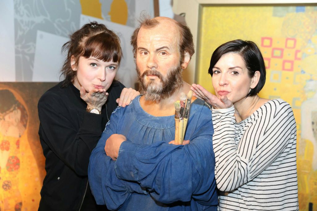 붓을 들고 있는 턱수염 왁스 피규어 옆에서 두 여성이 피규어의 얼굴에 입맞춤을 흉내 내며 포즈를 취하고 있습니다.