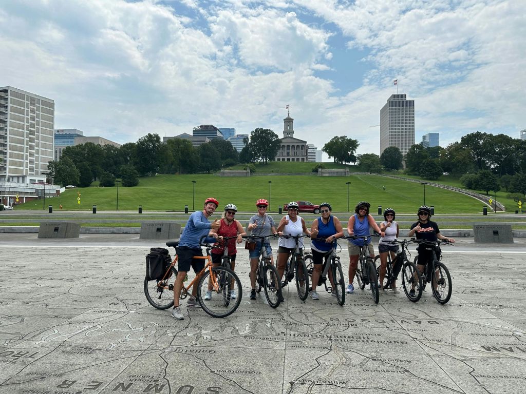 건물과 도시의 스카이라인을 배경으로 푸른 언덕 앞에서 포즈를 취하고 있는 한 무리의 사람들.