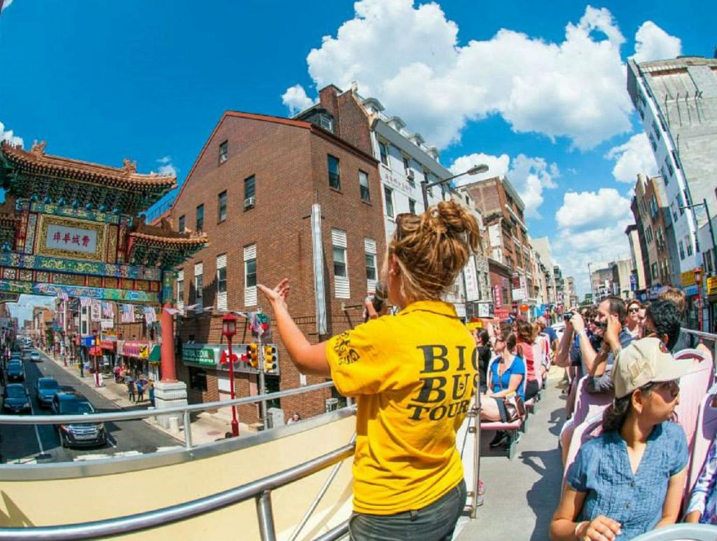 노란색 "빅 버스 투어" 셔츠를 입은 투어 가이드가 오픈탑 버스를 타고 도시 건물과 푸른 하늘을 배경으로 관광객과 이야기를 나눕니다.