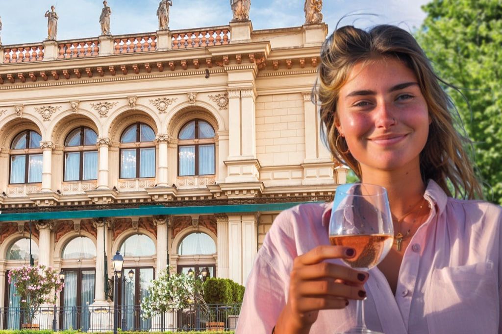 Junge Frau mit Weinglas lächelt vor dem prachtvollen Kursalon Hübner in Wien bei sonnigem Wetter.