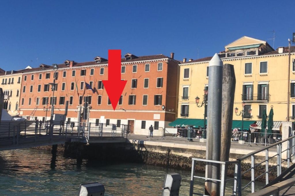 수변 산책로를 걷는 사람들과 함께 운하를 따라 건물을 짓습니다. 커다란 빨간색 화살표가 건물 중 한 곳을 가리킵니다.