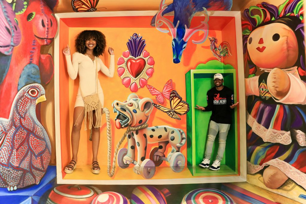 한 여성이 다양한 동물 모티브와 생동감 넘치는 나비가 있는 다채롭고 초현실주의적인 벽화 안에 서 있습니다.