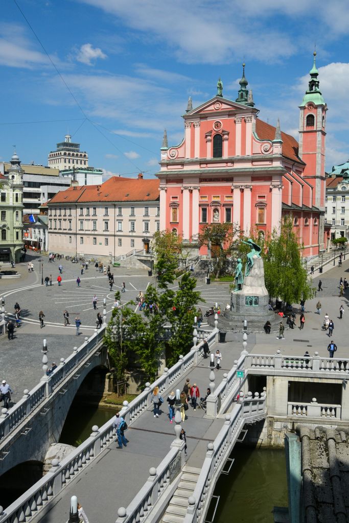 산책하는 사람들이 있는 번화한 도시 광장, 분홍색 바로크 양식의 교회, 다리, 중앙에 동상이 있습니다.