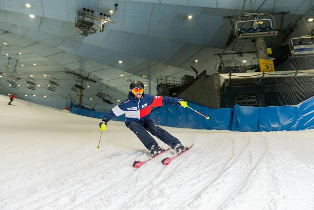스키 리프트를 배경으로 실내 슬로프에서 빨간색, 흰색, 파란색 재킷과 검은색 헬멧 스키를 입은 스키어.