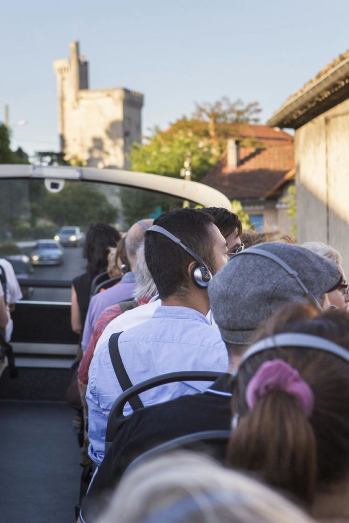 건물과 멀리 떨어진 타워가 있는 마을을 통과하는 오픈탑 투어 버스에 앉아 헤드폰을 착용한 사람들.