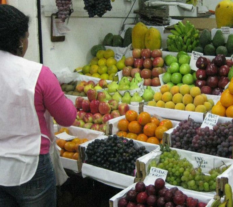 분홍색 셔츠와 흰색 앞치마를 입은 사람이 시장 노점에서 사과, 오렌지, 포도 등 다양한 과일을 배열하고 있습니다.