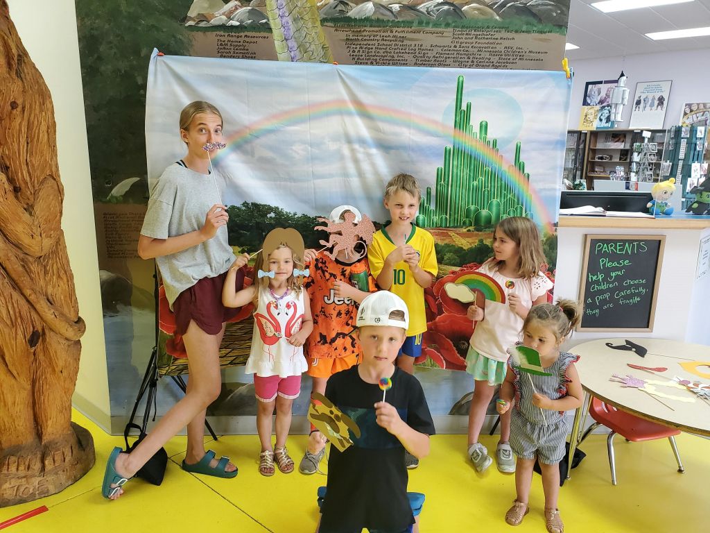 여섯 명의 어린이가 무지개와 에메랄드 시티가 있는 오즈 테마의 배경 앞에서 소품을 들고 포즈를 취하고 있습니다.