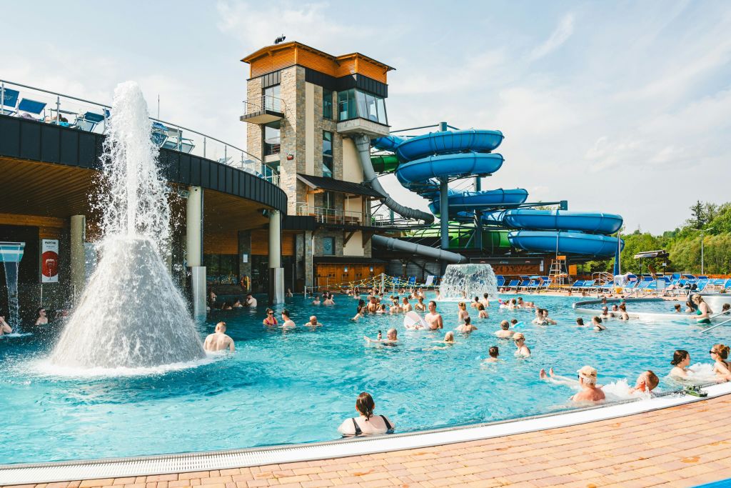 워터 슬라이드, 분수, 맑은 하늘 아래 복층 건물이 있는 붐비는 수영장 공간.