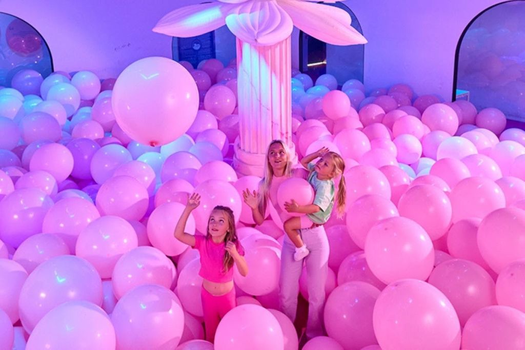 한 여성이 분홍색 풍선으로 둘러싸인 아이를 안고 있으며, 옆에 다른 아이가 커다란 기둥이 있는 분홍색 조명 방에 있습니다.