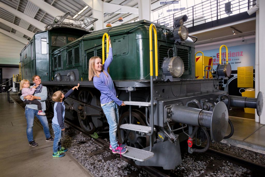 세 자녀를 둔 한 여성이 박물관에서 오래된 녹색 열차를 탐험하고 있습니다. 한 어린이는 계단을 오르며, 다른 어린이는 기차를 가리킵니다.