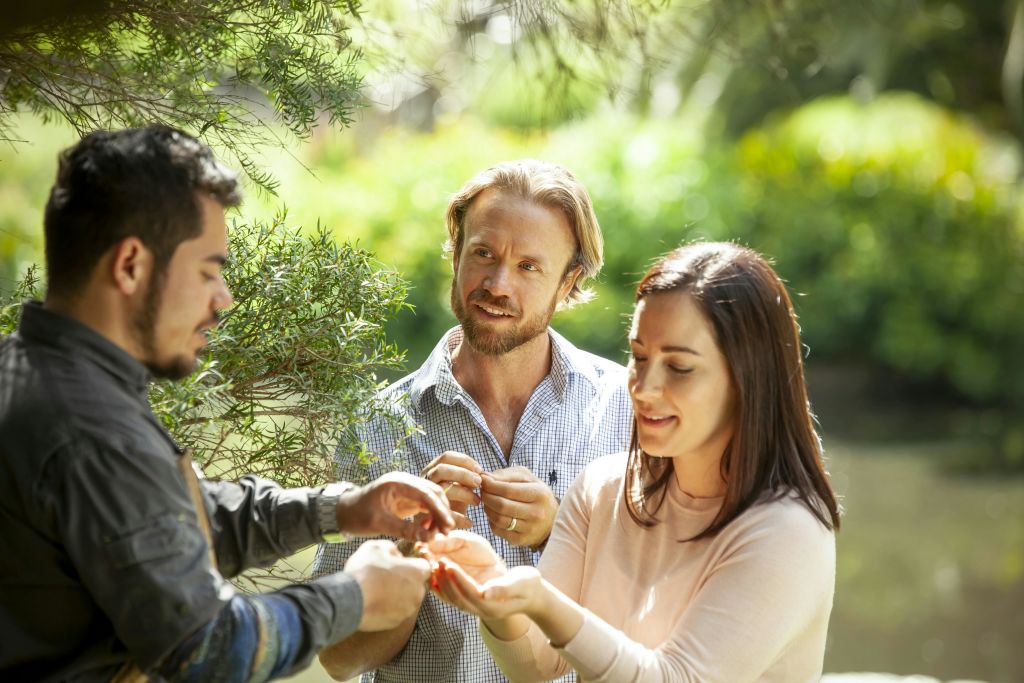 녹지대 근처에 야외에 서 있는 세 사람. 한 남자가 웃고 있는 여성에게 작은 물건을 건네고 있고, 다른 남자는 이를 바라보고 있습니다.