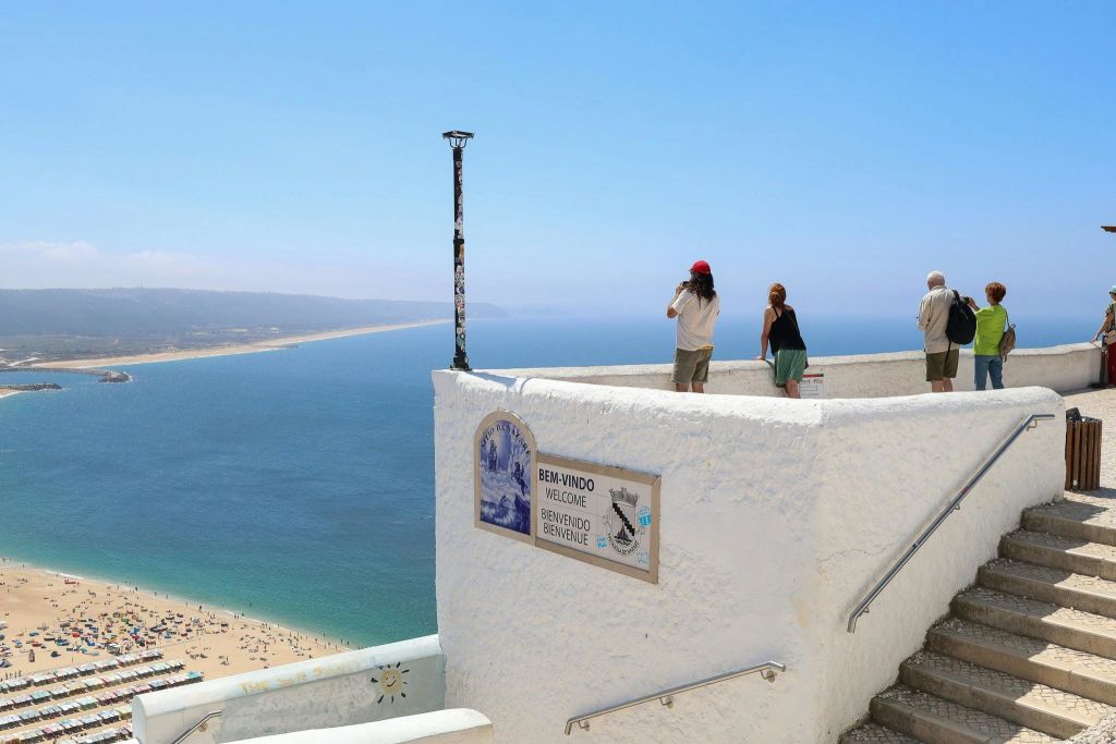 해변과 바다가 내려다보이는 하얀 벽으로 둘러싸인 전망대에 서 있는 사람들. 다국어로 된 표지판이 방문객을 맞이합니다.