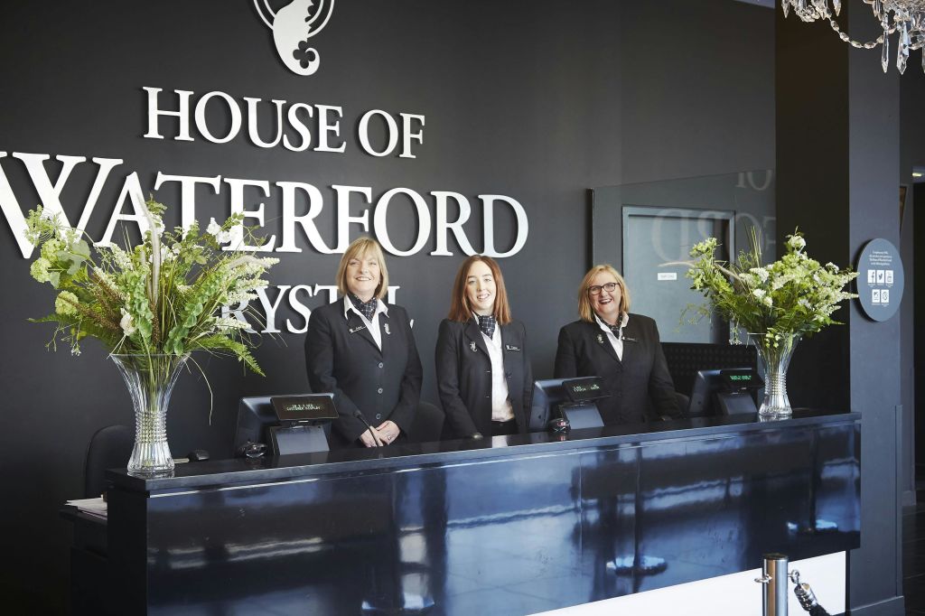검은색 유니폼을 입은 세 명의 여성이 House of Waterford Crystal 리셉션 구역의 프런트 데스크 뒤에 서 있습니다.