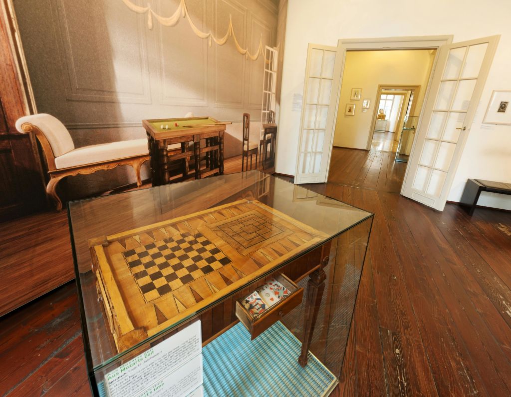 나무 바닥이 있는 방에는 유리 케이스 안에 체스 테이블이 있습니다. 개방된 출입구를 통해 인접한 방이 보입니다.