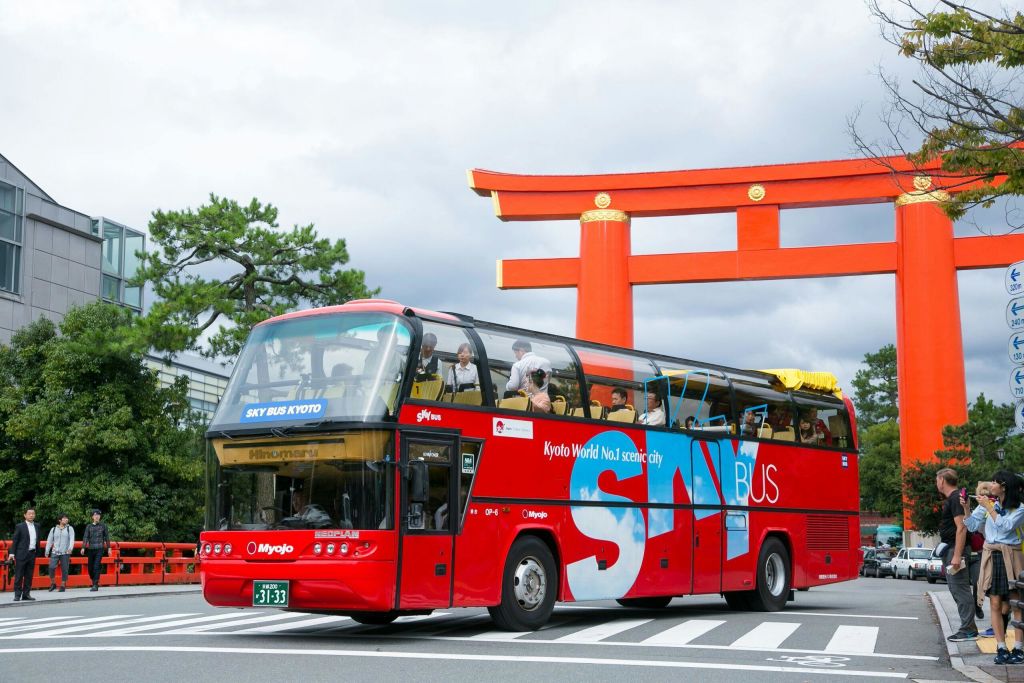 'SKY BUS KYOTO' 라고 표시된 빨간색 2층 관광 버스가 커다란 빨간색 토리이 게이트 아래 흐린 하늘을 배경으로 지나갑니다.