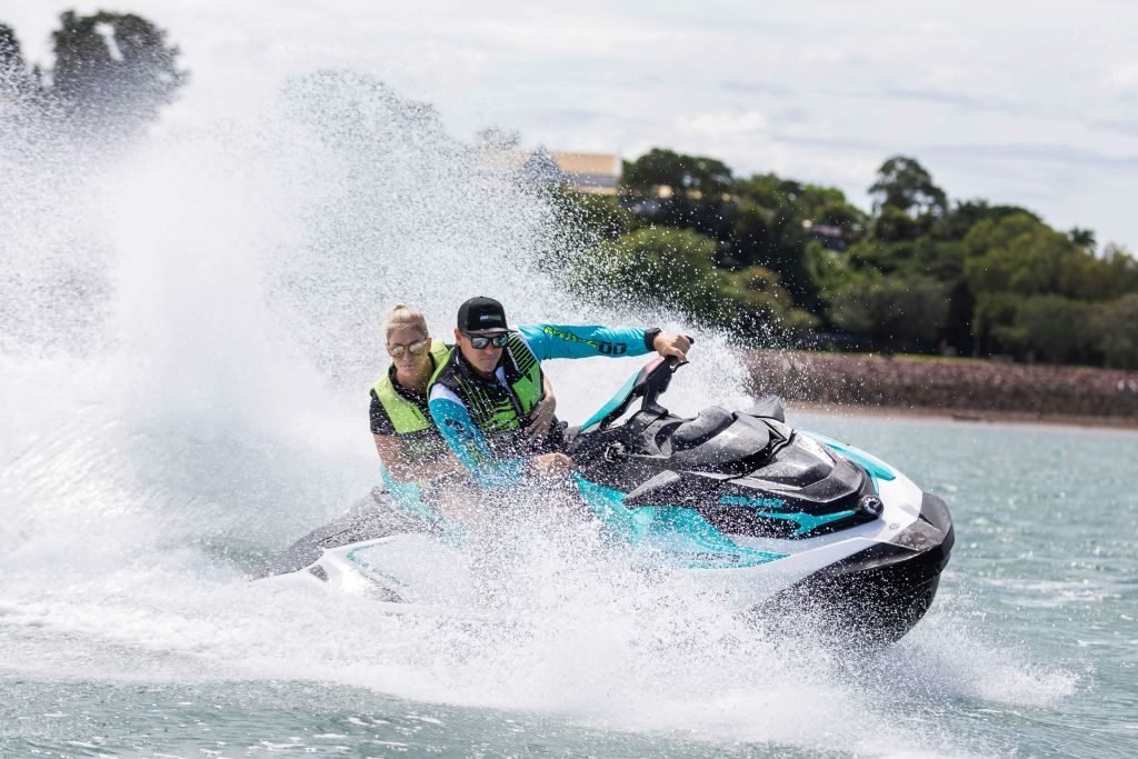 구명조끼와 고글을 착용한 두 사람이 제트 스키를 타고 물 위를 고속으로 달리며 커다란 물보라를 일으킵니다. 나무는 배경에 있습니다.