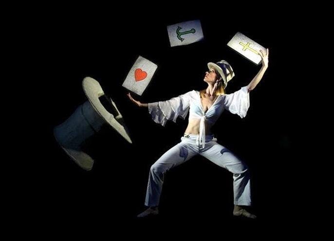 흰색 의상과 모자를 쓴 사람이 어두운 배경에 하트, 닻, 십자가를 상징하는 커다란 카드를 저글링합니다.