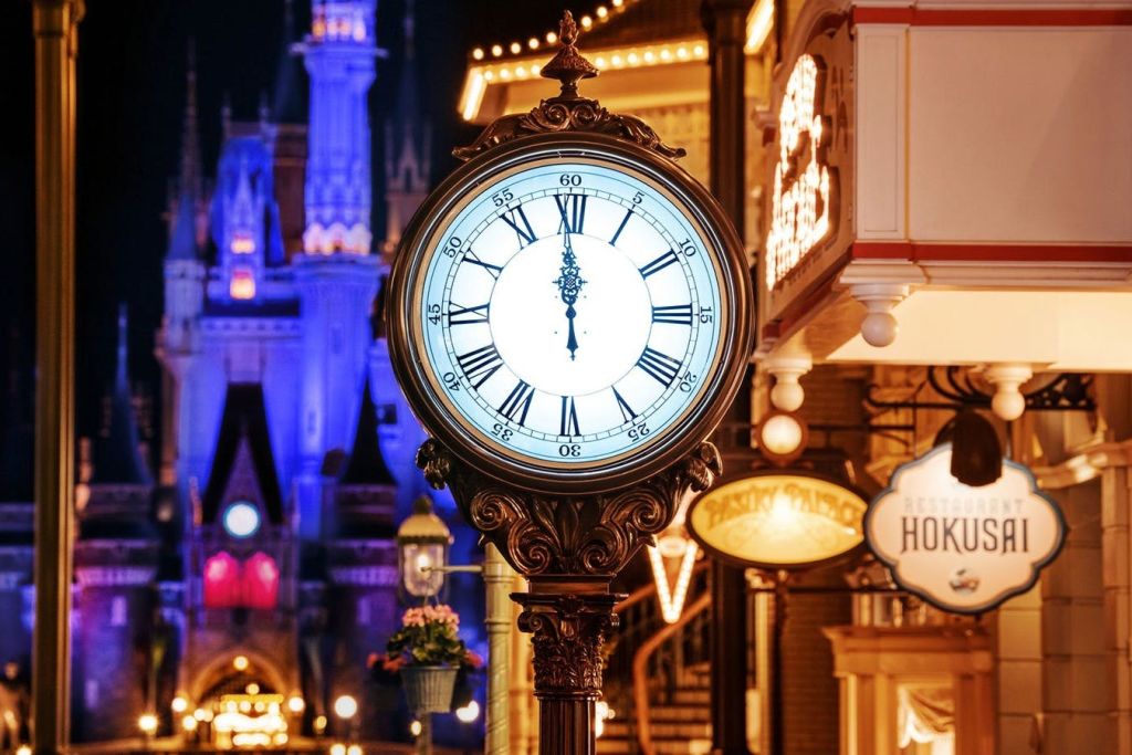 11시 55분에 로마 숫자가 새겨진 화려한 거리 시계, 조명이 켜진 건물, 파란색과 보라색으로 배경이 된 저 멀리 보이는 성.
