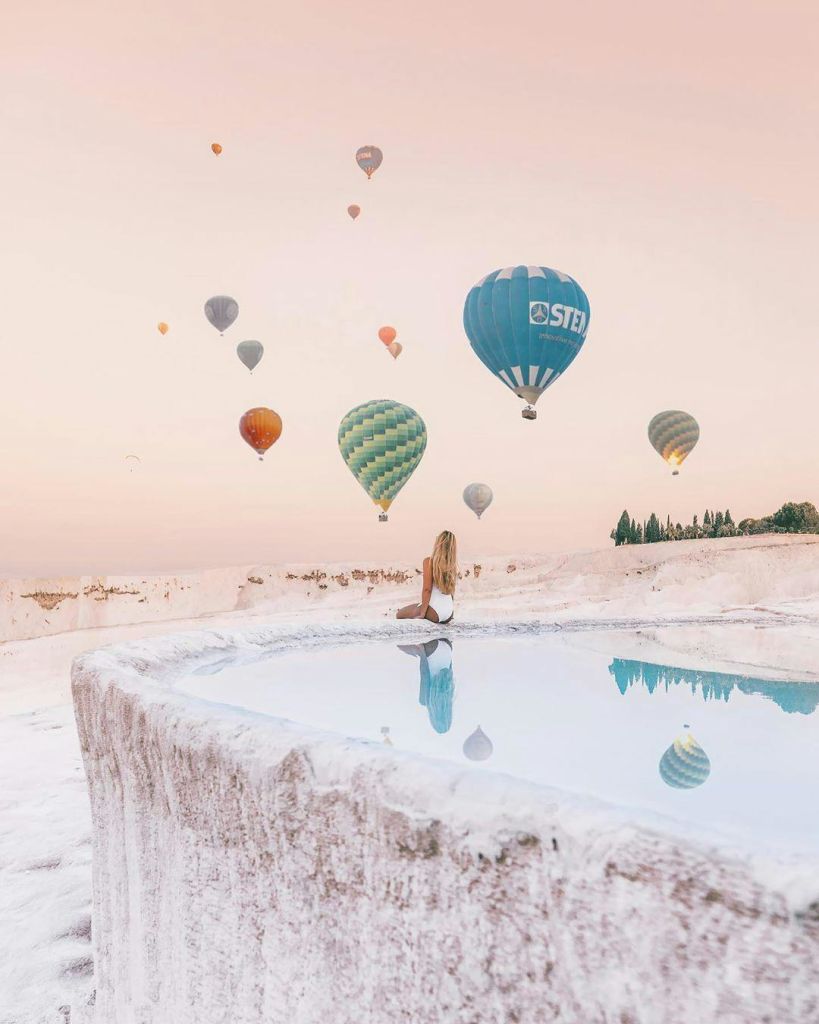 수영복을 입은 사람이 반사 수영장에 앉아 화려한 열기구가 파스텔 하늘에 떠 있는 것을 지켜보고 있습니다.