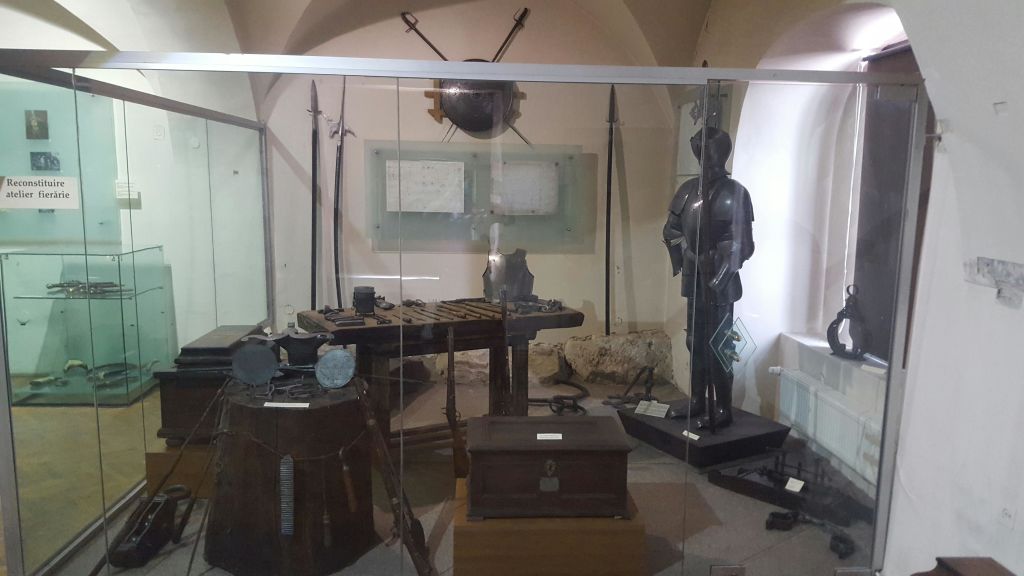 역사적인 갑옷, 무기, 나무 상자 및 문서를 전시하는 박물관 전시물입니다. 갑옷을 입은 마네킹이 오른쪽에 서 있습니다.