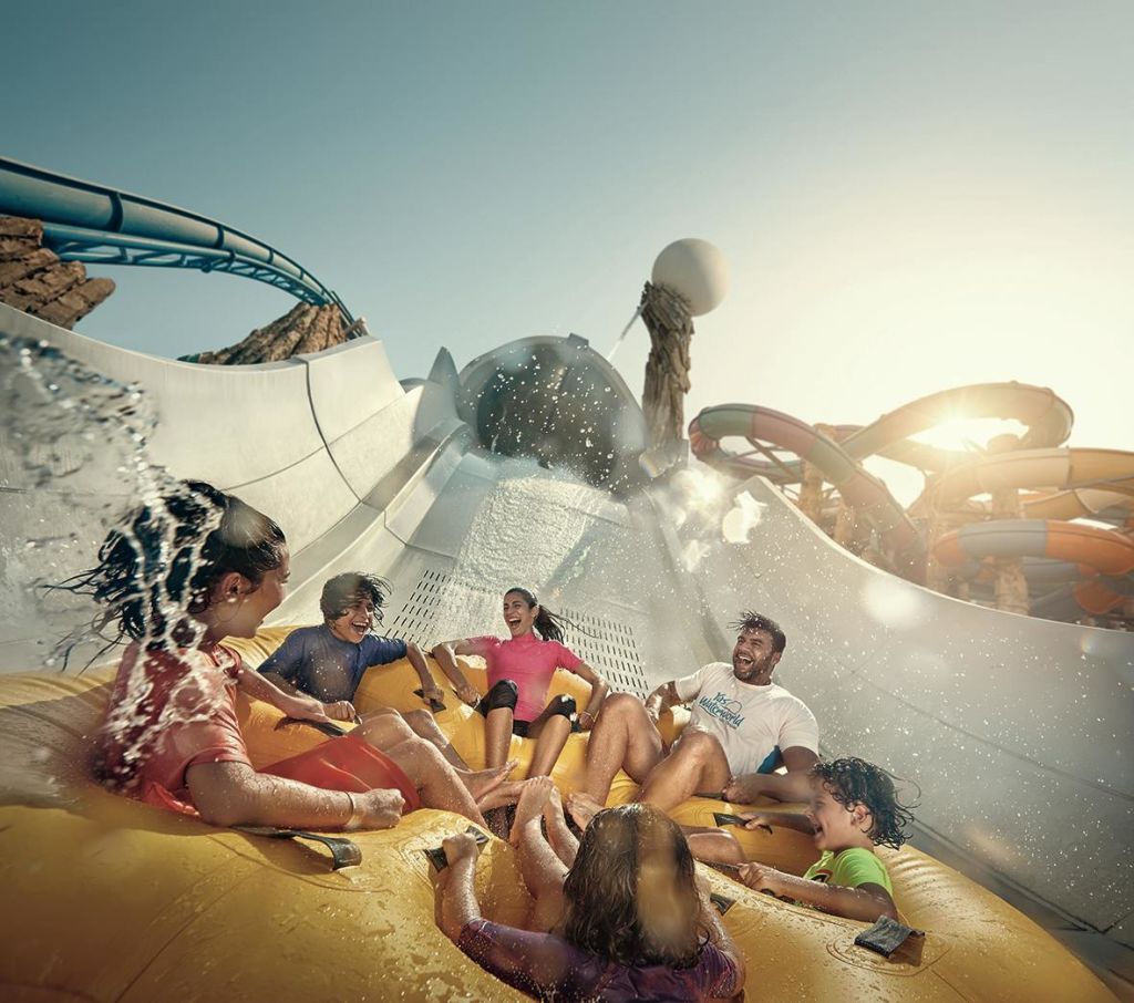6명의 일행이 커다란 팽창식 뗏목을 타고 워터 슬라이드를 타며 주변의 물을 튀기고 있습니다.