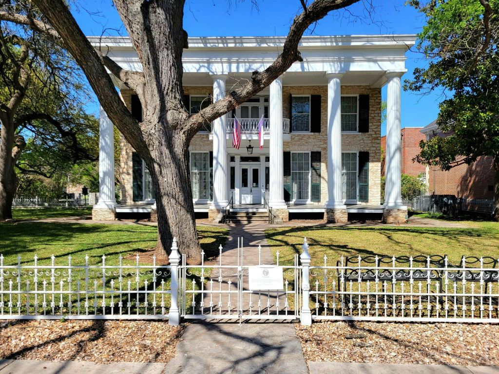 흰색 기둥이 높고 철제 울타리가 있으며 입구에 미국 국기 두 개가 있는 2층짜리 대형 주택입니다.