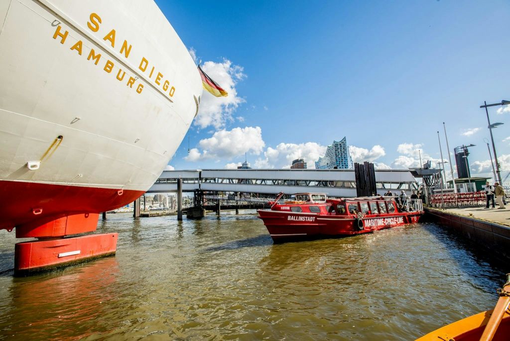 "샌디에고" 라고 표시된 대형 흰색 선미와 "발린슈타트" 라고 표시된 빨간색 투어 보트가 도시 풍경과 엘프필하모니를 배경으로 있습니다.