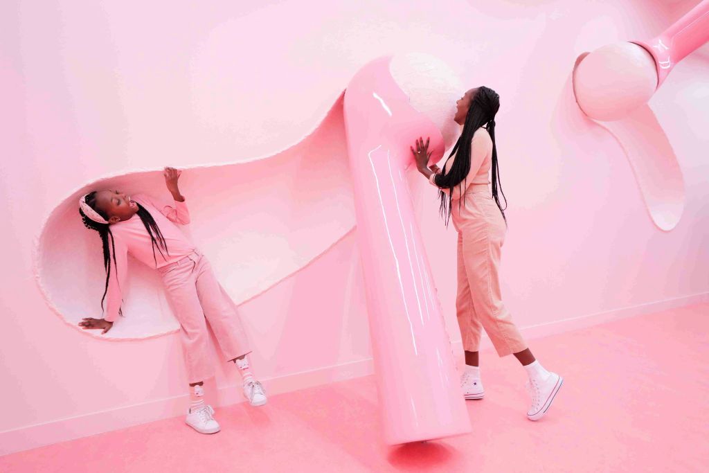 분홍색 의상을 입은 두 사람이 단색 분홍색 방에서 커다란 분홍색과 흰색 추상 구조물과 상호 작용합니다.