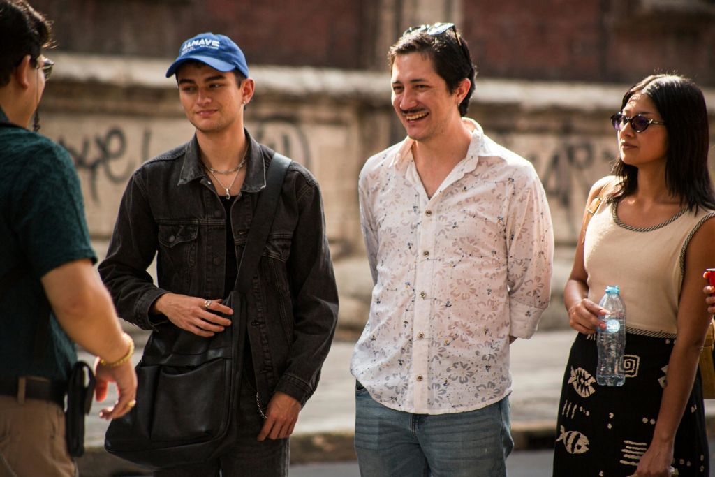 세 사람이 야외에 서서 대화를 나누고 있습니다. 한 명은 파란색 모자를 쓰고, 다른 한 명은 흰색 셔츠를 입고, 배경에는 낙서가 보입니다.