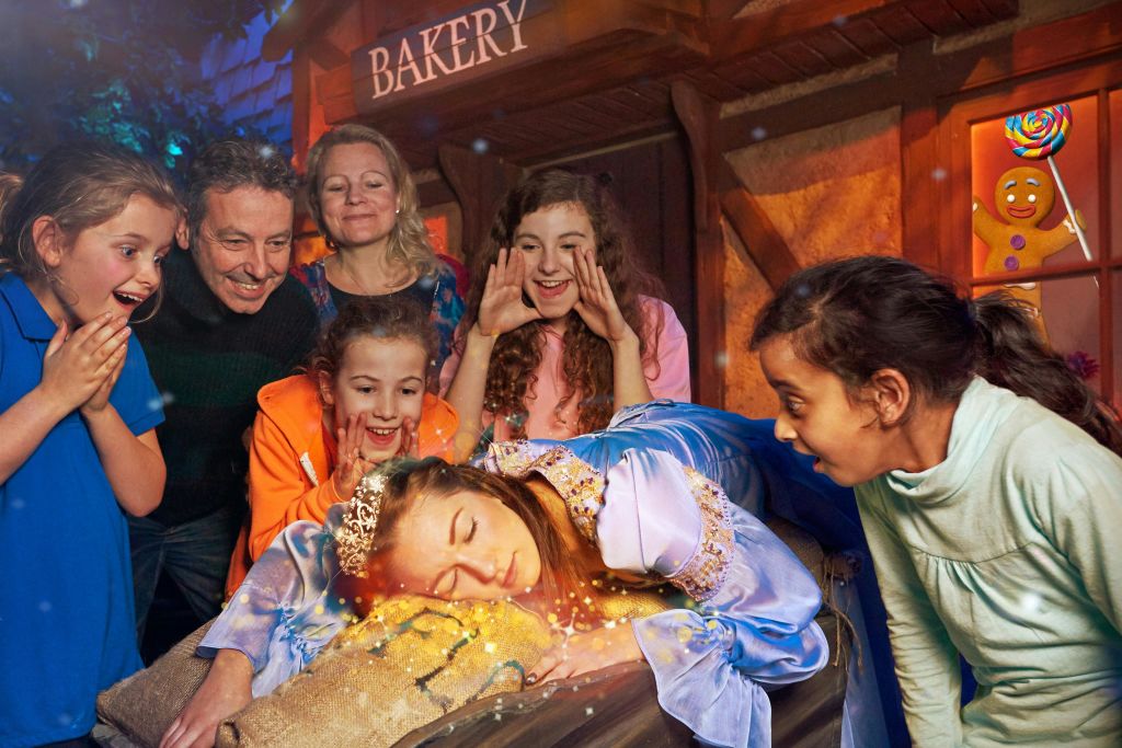 소박한 침대 위에 누워 있는 잠자는 숲속의 미녀로 분장한 한 사람 주위에 여섯 명이 모였다. 그들 뒤에는 "베이커리" 표지판이 보입니다.