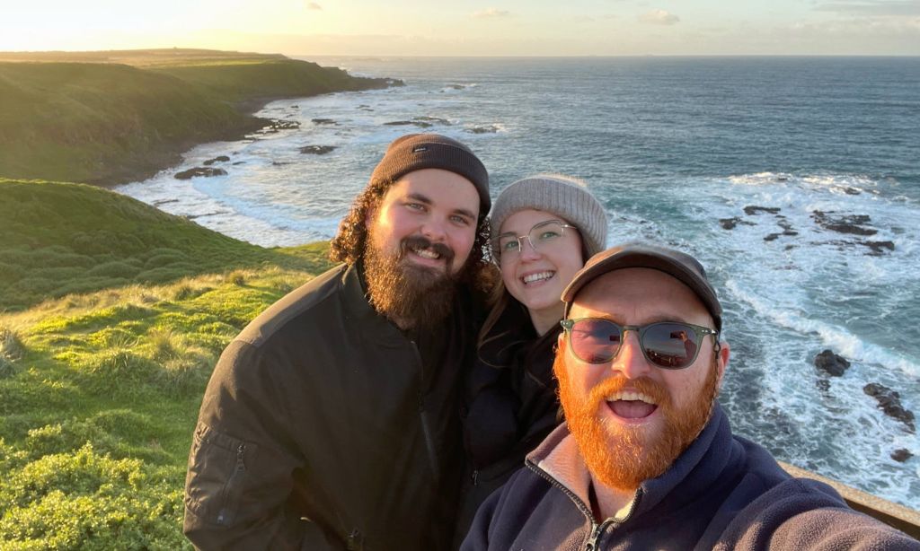 부분적으로 흐린 하늘 아래 절벽과 바다 파도가 있는 경치 좋은 해안가에서 웃고 있는 세 사람.