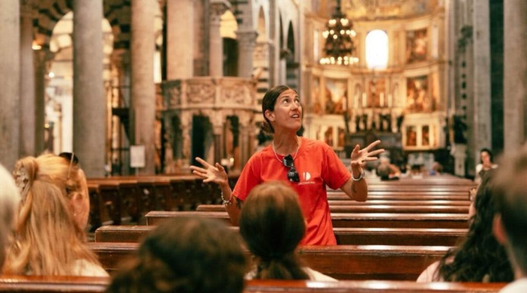 피사 대성당 앞에 서 있는 한 무리의 관광객은 맑은 하늘 아래에서 막대기에 깃발을 들고 있습니다.