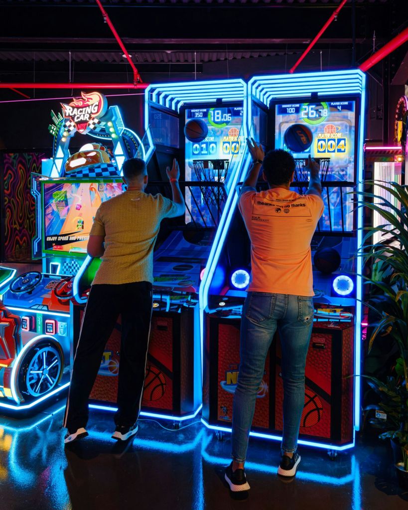 네온 조명과 레이싱 게임을 배경으로 밝게 빛나는 아케이드에서 농구 아케이드 게임을 즐기는 두 사람.