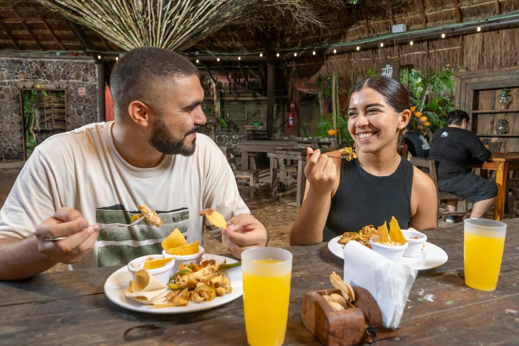 나무 가구와 줄 조명이 있는 소박한 야외 레스토랑에서 두 사람이 함께 미소 짓고 식사를 합니다.